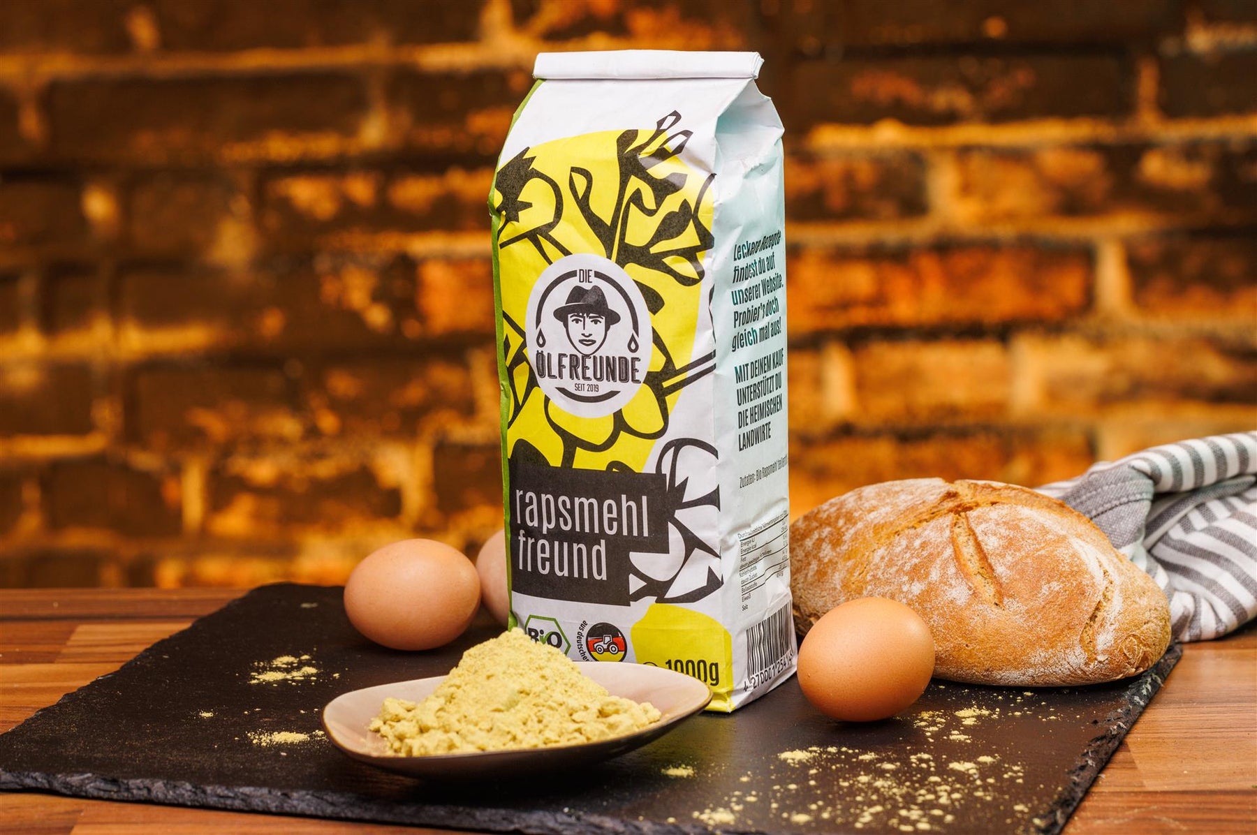 Eine Tüte mit 1000g Bio-Rapsmehl der Marke Die Ölfreunde, neben einer Schale mit Rapsmehl, Eiern und Brot.
