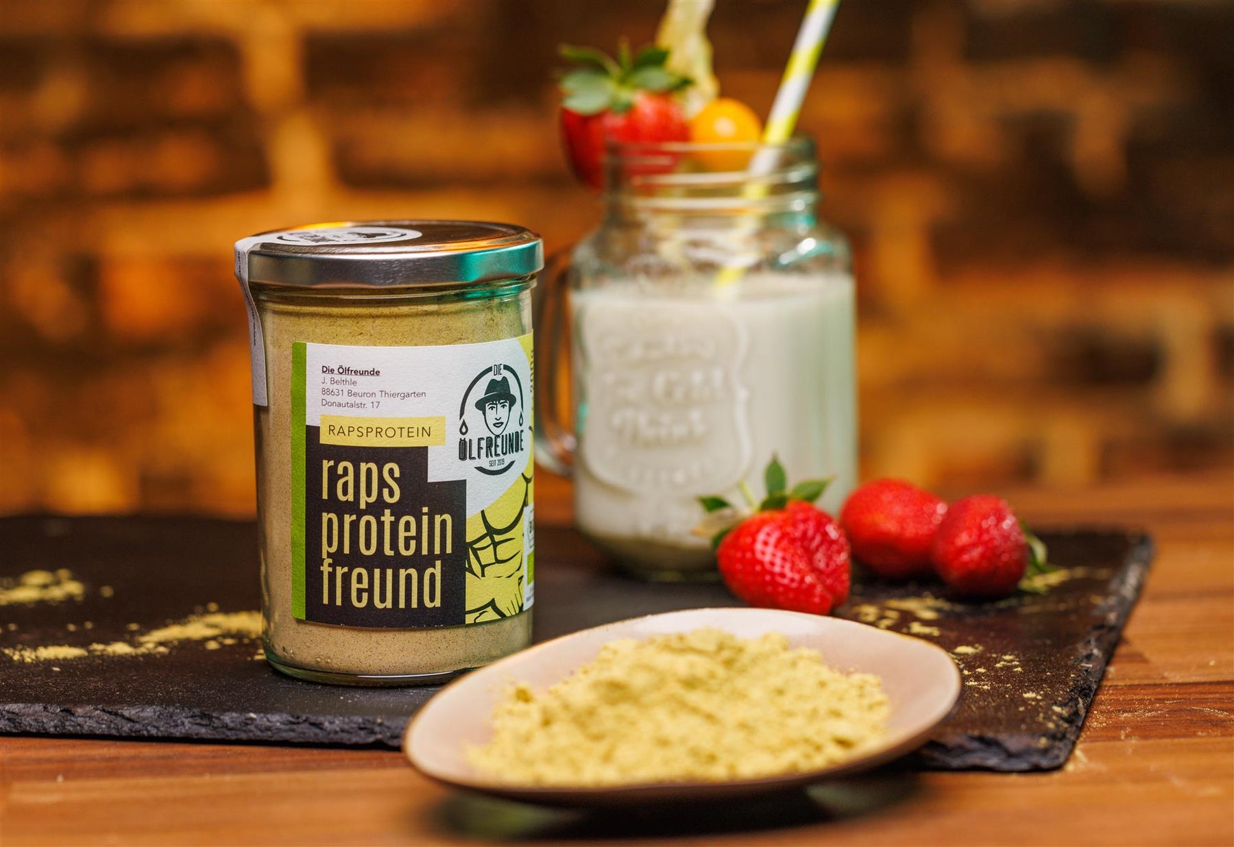 Ein Glas mit 200g Bio-Rapsprotein der Marke Die Ölfreunde, daneben eine Schale mit Rapsprotein, Erdbeeren und im Hintergrund ein Milchhake.