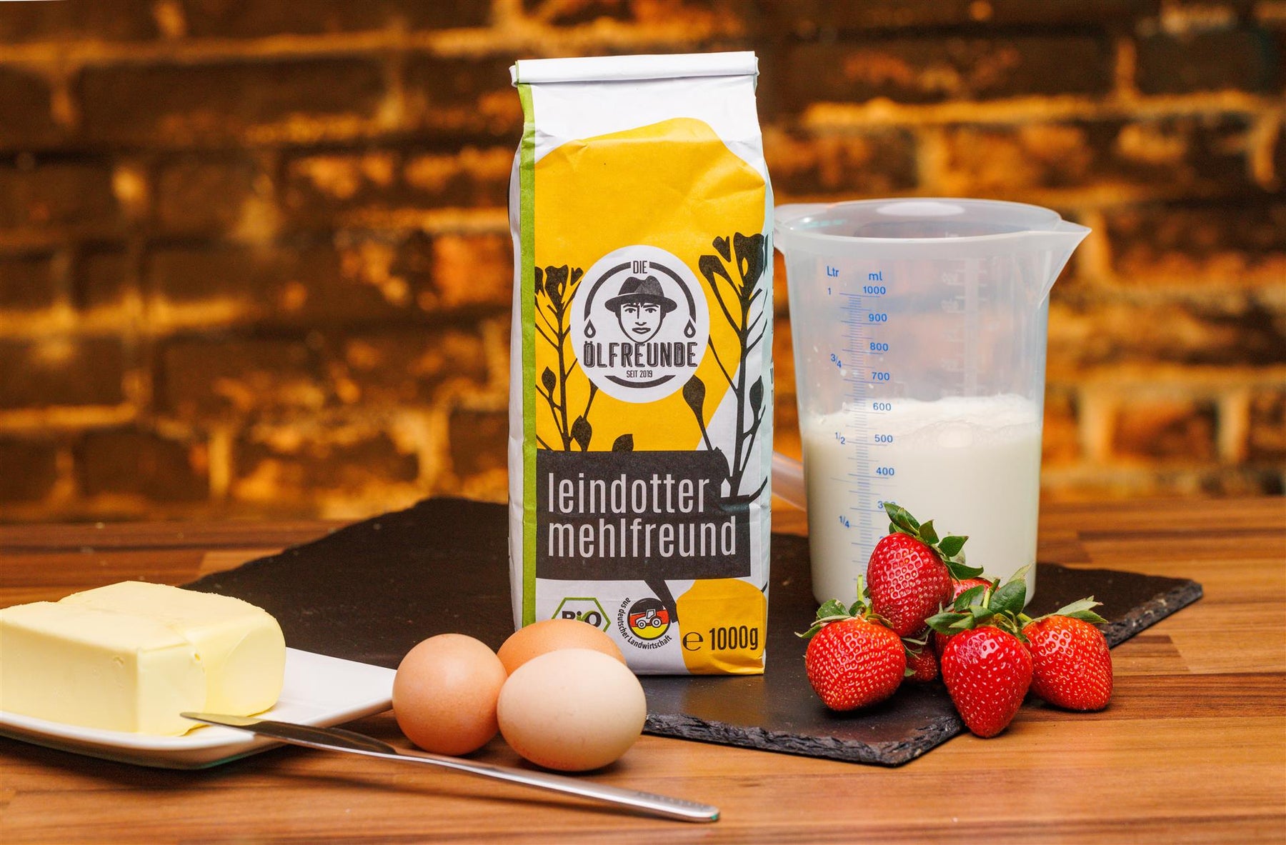 Im Zentrum steht eine Packung Leindottermehl der Marke Die Ölfreunde, rechts daneben Erdbeeren und ein Behälter mit Milch. Links liegen Eier, ein Messer und ein Teller mit einem Block Butter