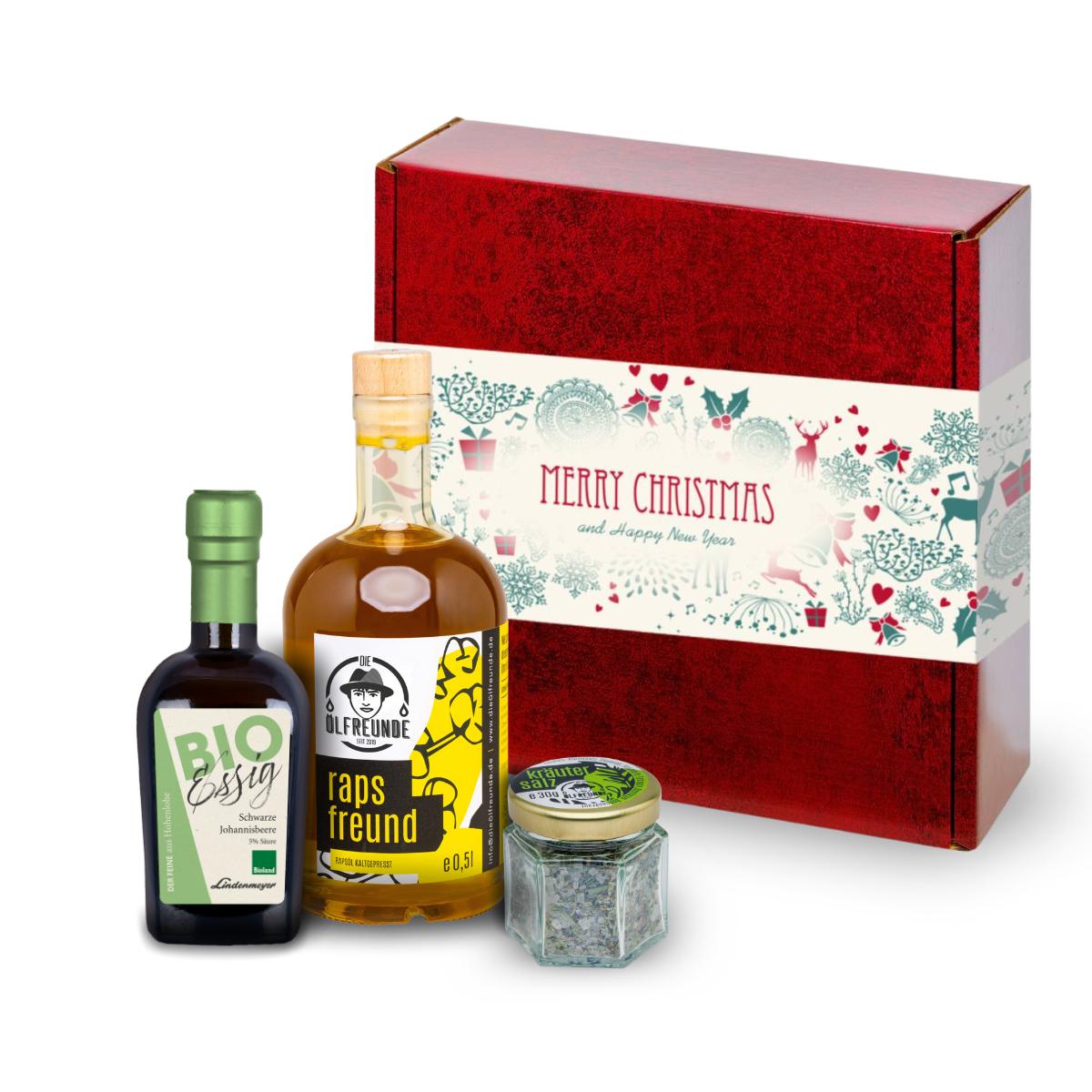 Eine Flasche Rapsöl und ein Glas Kräutersalz der Marke Die Ölfreunde und eine Flasche Bio-Essig Schwarze Johannisbeere der Marke Lindenmeyer, vor einer Weihnachts-Geschenkeverpackung.