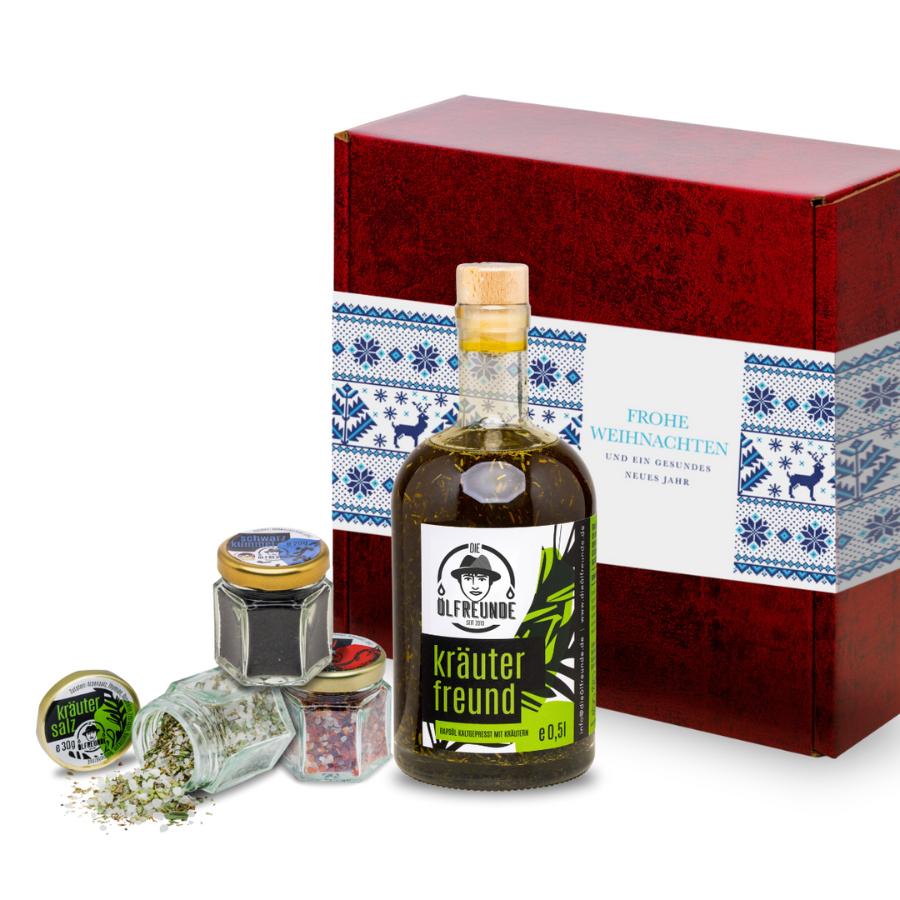 Eine Flasche Kräuteröl, ein Glas Schwarzkümmel, Kräutersalz und Chillisalz der Marke Die Ölfreunde, vor einer Weihnachts-Geschenkeverpackung