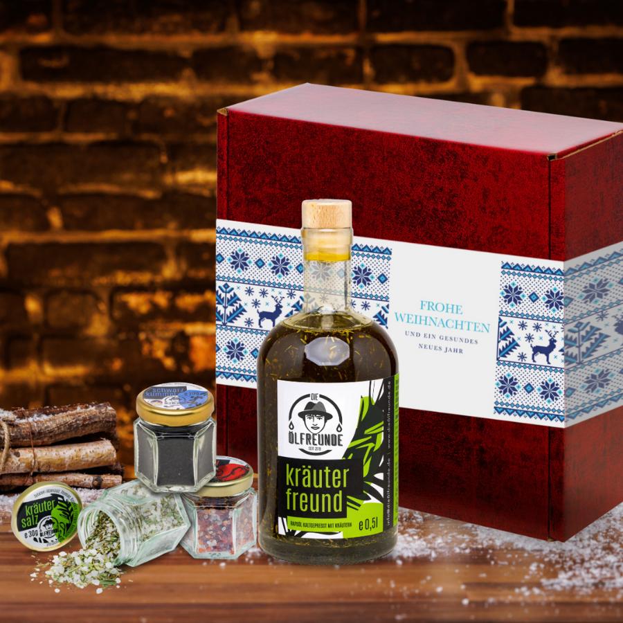 Eine Flasche Kräuteröl, ein Glas Schwarzkümmel, Kräutersalz und Chillisalz der Marke Die Ölfreunde, vor einer Weihnachts-Geschenkeverpackung und neben einem Bündel Holz.