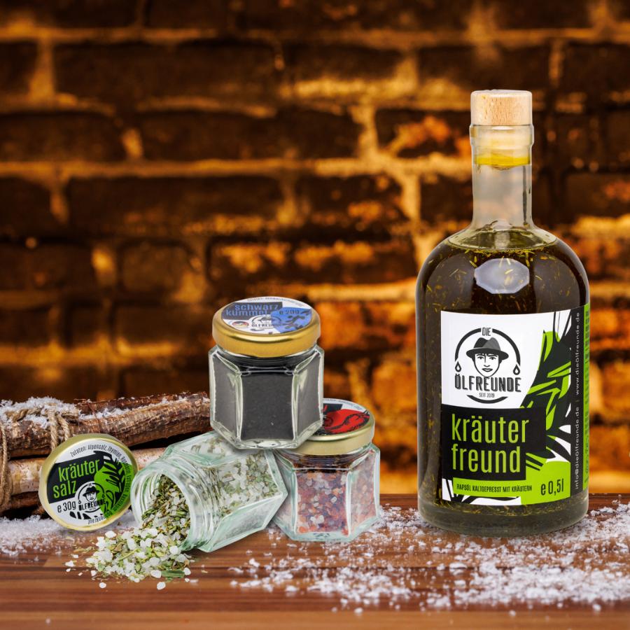 Eine Flasche Kräuteröl, ein Glas Schwarzkümmel, Kräutersalz und Chillisalz der Marke Die Ölfreunde, neben einem Bündel Holz.