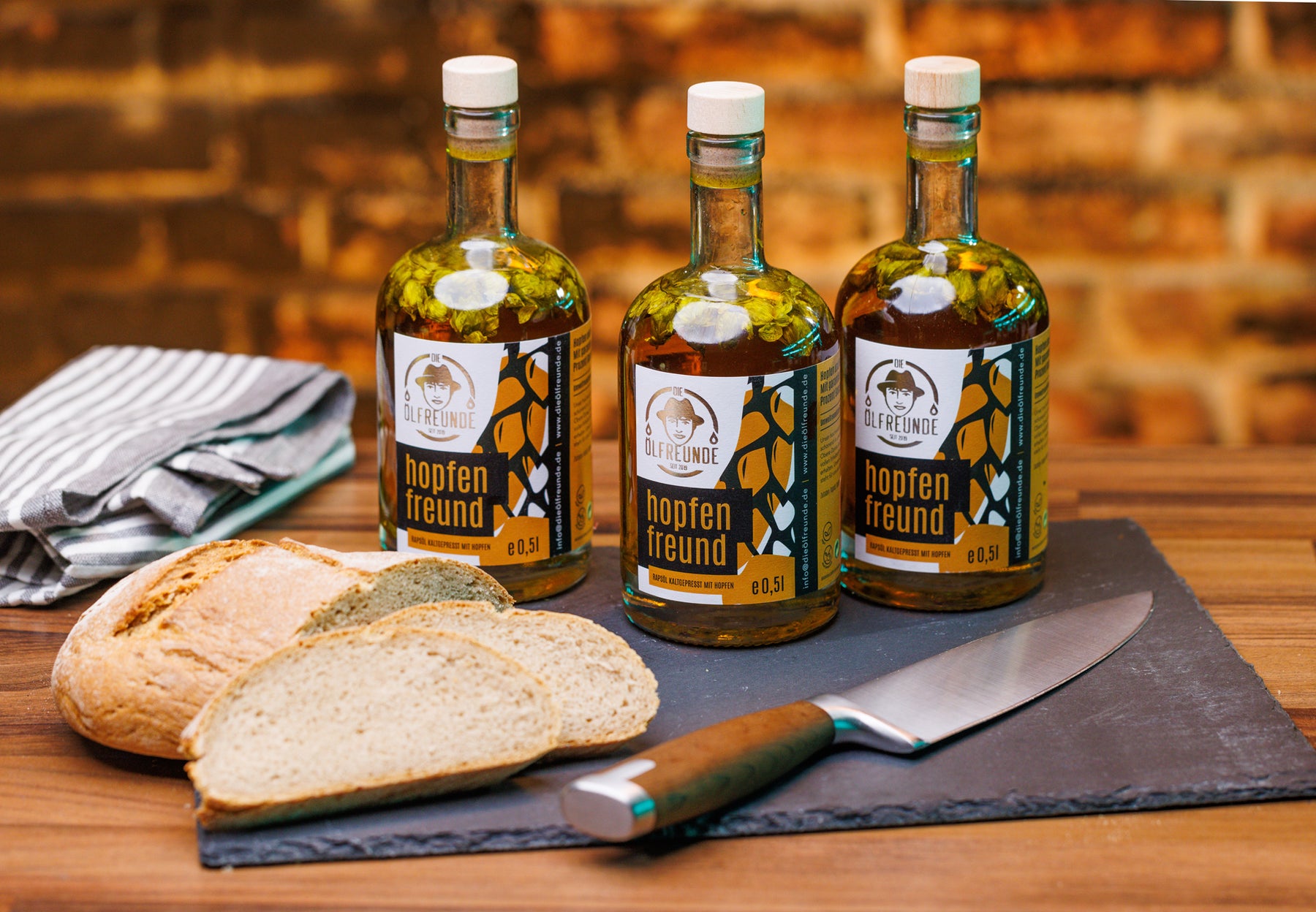 Drei Flaschen kaltgepresstes Rapsöl mit Hopfen, der Marke Die Ölfreunde, umgeben von einem Geschirrtuch, Brot und einem Messer