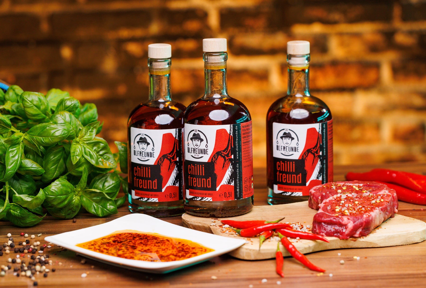 Drei Flaschen kaltgepresstes Chilli-Öl mit crispy Chilliflocken, von Marke Die Ölfreunde, daneben ein rohes, gewürztes Steak, frischer Basilikum, frische Chillis und ein Teller mit Chilliöl.
