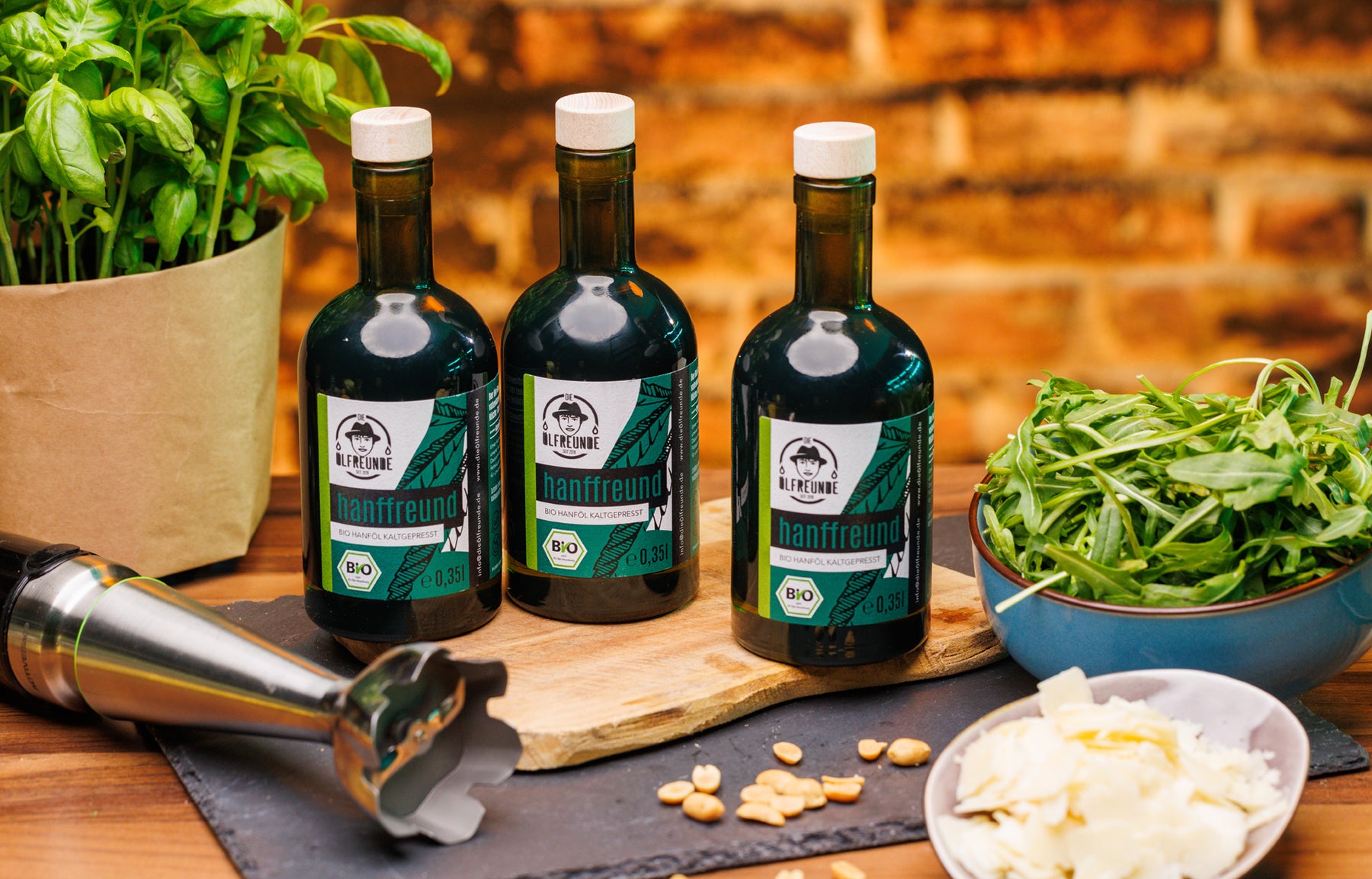 Drei Flaschen Hanföl der Marke Die Ölfreunde, auf einem Holzbrett, neben einer Schale mit Rucola, Parmesan, Pinienkerne und einen Pürieraufsatz.