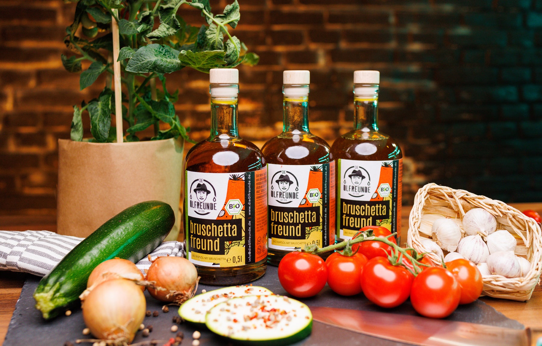 Drei Flaschen Bruschetta-Öl der Marke Die Ölfreunde, umgeben von frischem Gemüse und zwei gewürzten Zucchini-Scheiben.