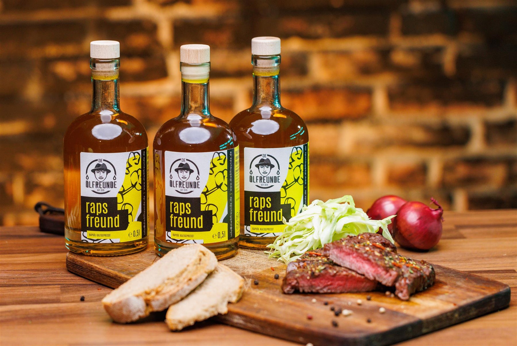 Drei Flaschen mit je 500ml Rapsöl der Marke Die Ölfreunde, auf einem Holzbrett, neben einem Stück Steak, Salat, Brot, Zwiebeln und einem Messer.