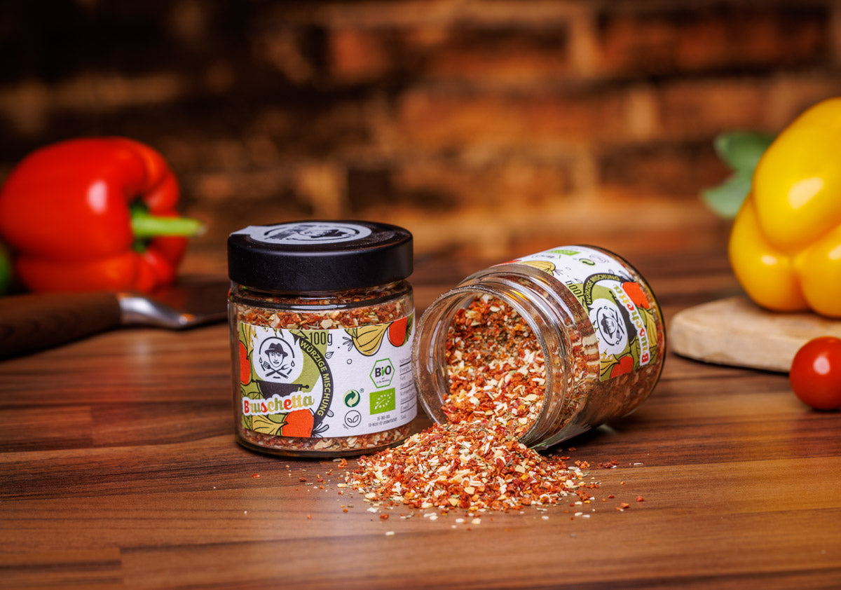 Zwei Gläser mit je 100g Bio-Bruschetta-Gewürzmischung der Marke Die Ölfreunde, auf rustikalem Holztisch: Ein Glas steht aufrecht, während das andere umgekippt ist und Gewürze verteilt, daneben frische Paprika.