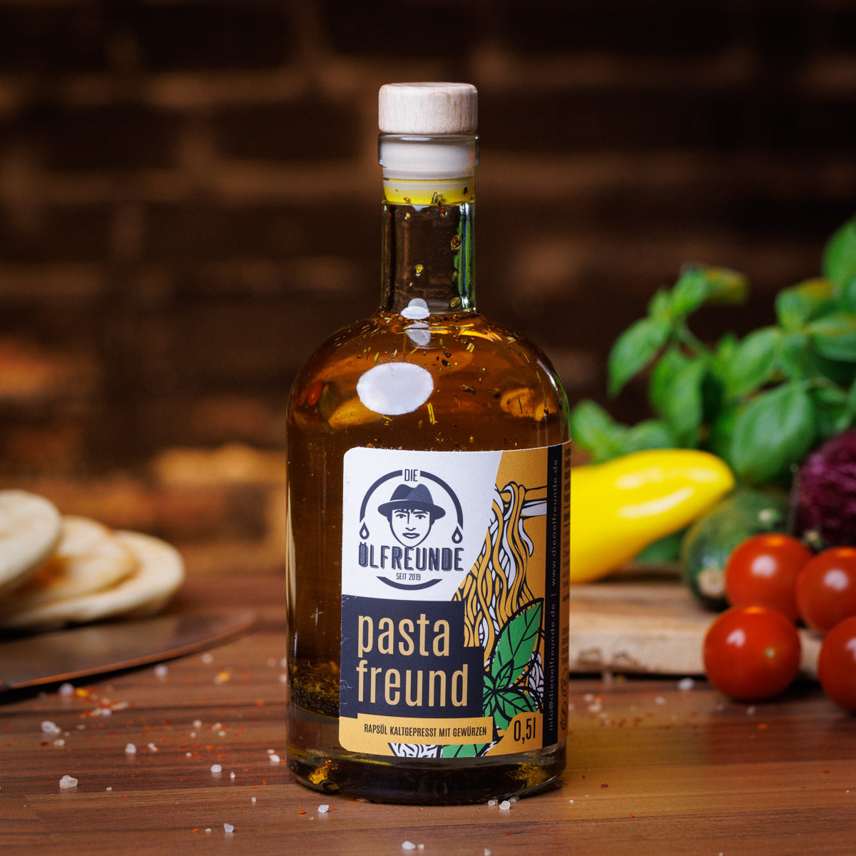 Eine Flasche Pasta-Öl der Marke Die Ölfreunde, auf holzigen Untergrund, neben frischem Gemüse