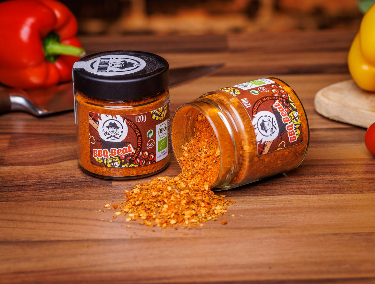 Zwei Gläser mit je 120g BBQ Beat Bio-Gewürzmischung der Marke Die Ölfreunde auf rustikalem Holztisch: Ein Glas steht aufrecht, während das andere umgekippt ist und Gewürze verteilt, daneben frische Paprika.