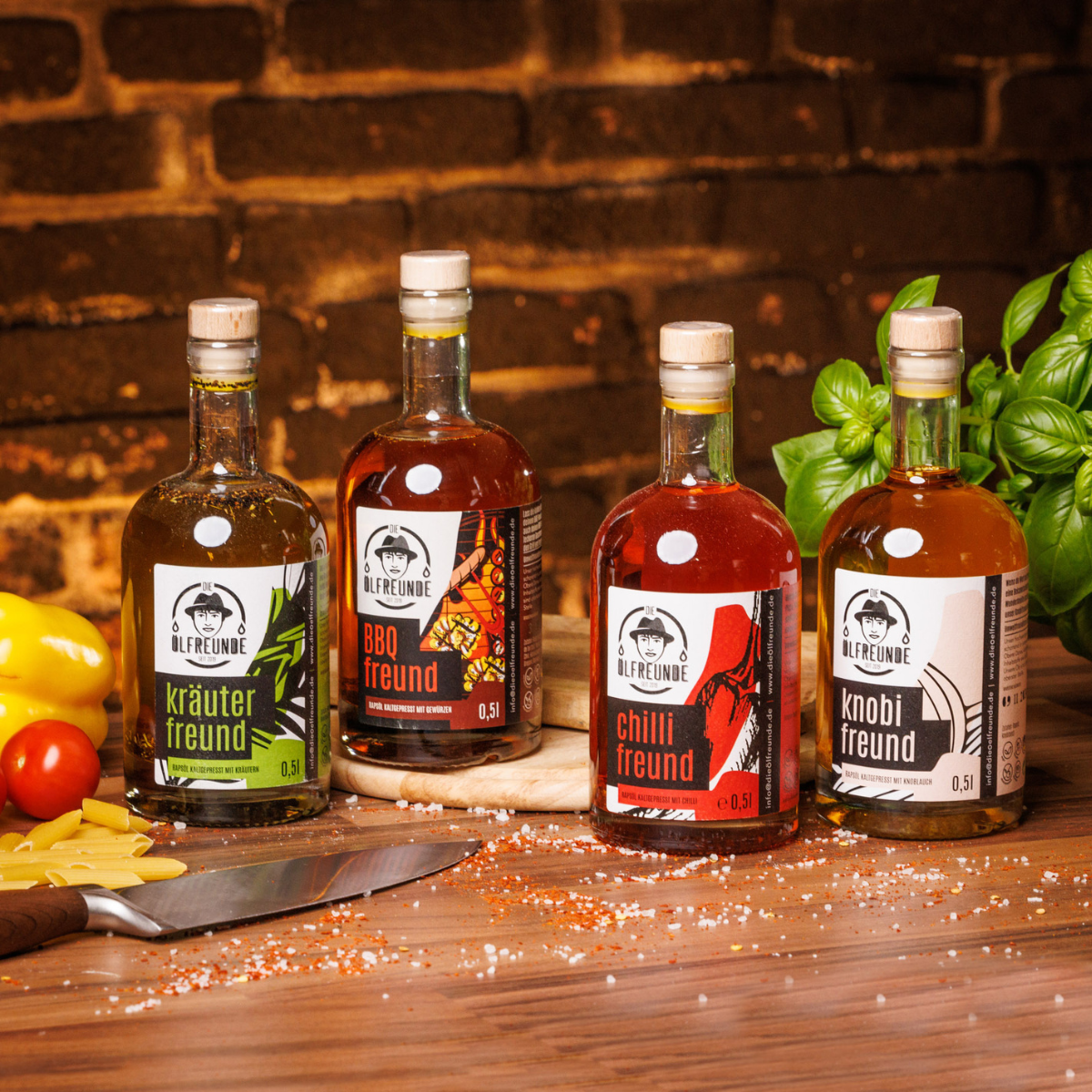Je eine Flasche Kräuteröl, BBQ-Öl, Chilliöl und Knoblauchöl der Marke Die Ölfreunde, auf einem Holzigen Untergrund, umgeben von frischem Gemüse, trockenen Nudeln, Gewürzen und einem Messer.