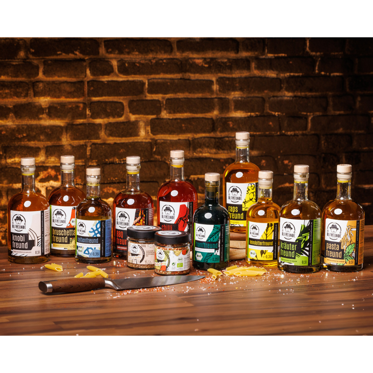 Je eine Flasche Knoblauchöl, Bruschetta-Öl, Leindotteröl mit Gewürzen, BBQ-Öl, Chilliöl, Hanföl, Rapsöl, Leindotteröl, Kräuteröl, Pasta-Öl, ein Glas Rauchsalz und ein Glas Bruschetta-Gewürzmischung der Marke Die Ölfreunde, auf Holzigen Untergrund, umgeben von einem Messer und Nudeln.
