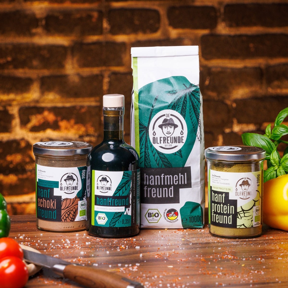 Ein Glas Trinkschokolade mit Hanf, ein Glas Hanfprotein, eine Flasche Hanföl und eine Tüte Hanfmehl der Marke Die Ölfreunde, auf holzigen Untergrund, umgeben von einem Messer, frischem Gemüse und Gewürzen.