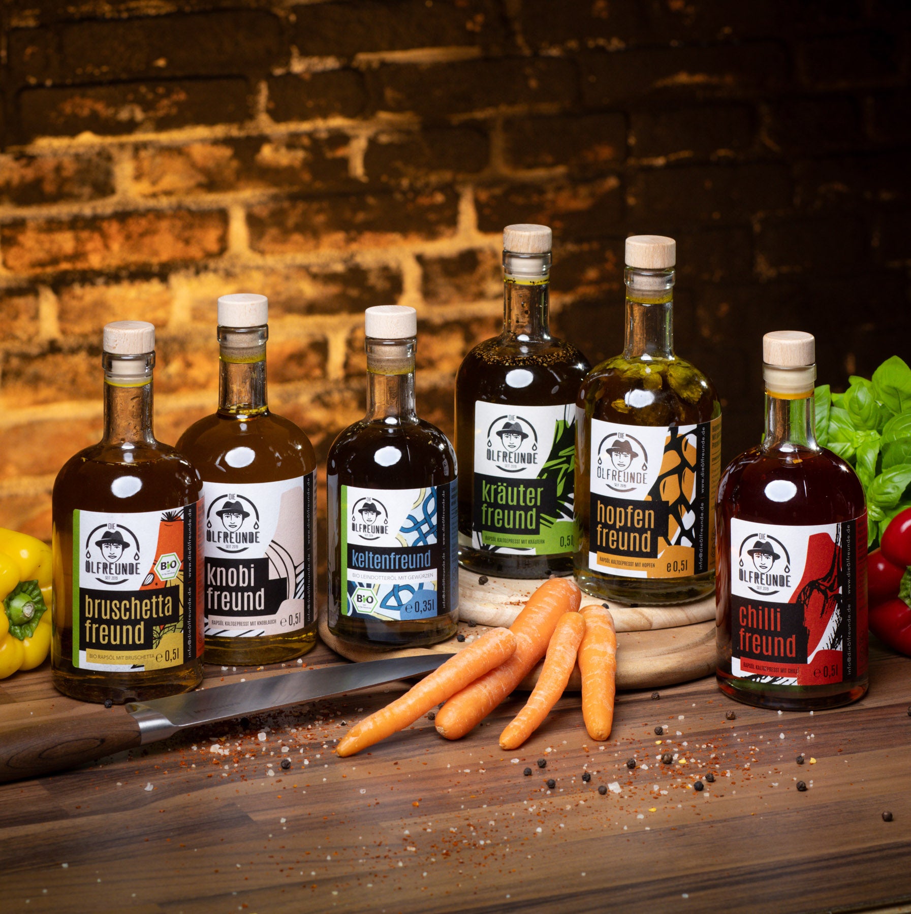 Je eine Flasche Bruschetta-Öl, Knoblauchöl, Leindotter-Öl mit Gewürzen, Kräuteröl, Hopfen-Öl und Chilliöl der Marke Die Ölfreunde, auf einem Holzigen Untergrund, umgeben von einem Messer, frischen Gemüse und Gewürzkörner.