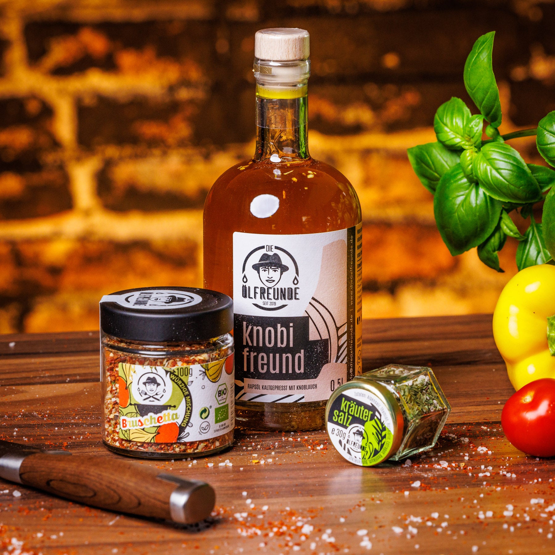 Eine Flasche Knoblauchöl, ein Glas Bruschetta-Gewürzmischung und ein Glas Kräutersalz der Marke Die Ölfreunde, auf einem holzigen Untergrund, neben frischem Gemüse.