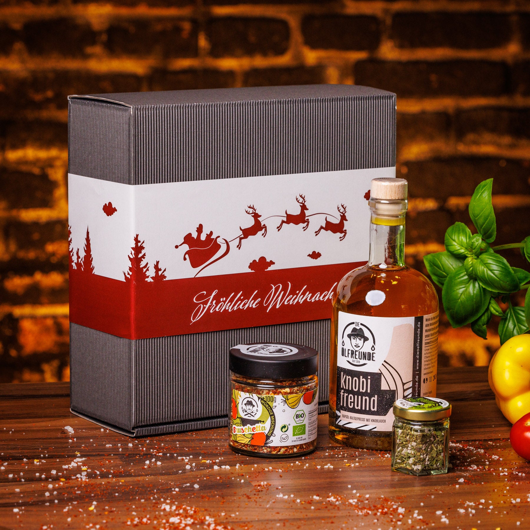 Eine Flasche Knoblauchöl, ein Glas Bruschetta-Gewürzmischung und ein Glas Kräutersalz der Marke Die Ölfreunde, vor einer Weihnachts-Geschenkverpackung, neben frischem Gemüse.