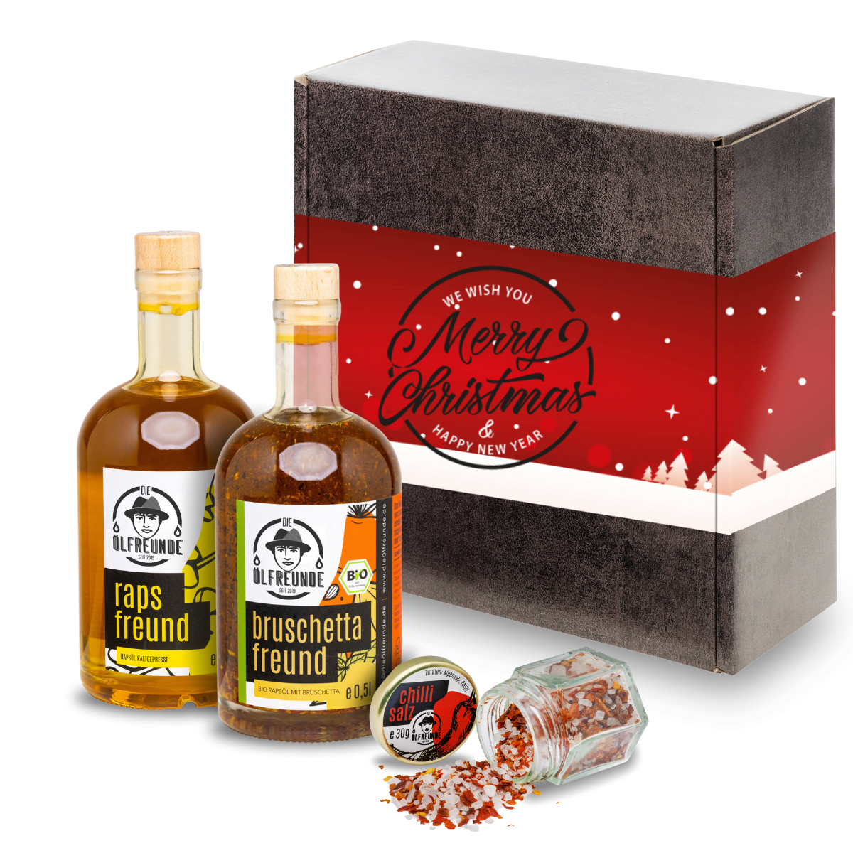 Eine Flasche Rapsöl, Bruschetta-Öl und ein Glas Chillisalz der Marke Die Ölfreunde, vor einer Weihnachts-Geschenkverpackung.