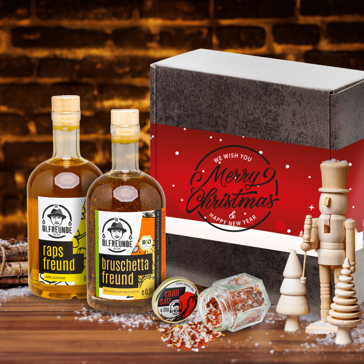 Ein Glas Chillisalz, eine Flasche Rapsöl und Bruschetta-Öl der Marke Die Ölfreunde vor einer Weihnachts-Geschenkverpackung, neben einem Bündel Holz und Weihnachtlicher Dekoration.