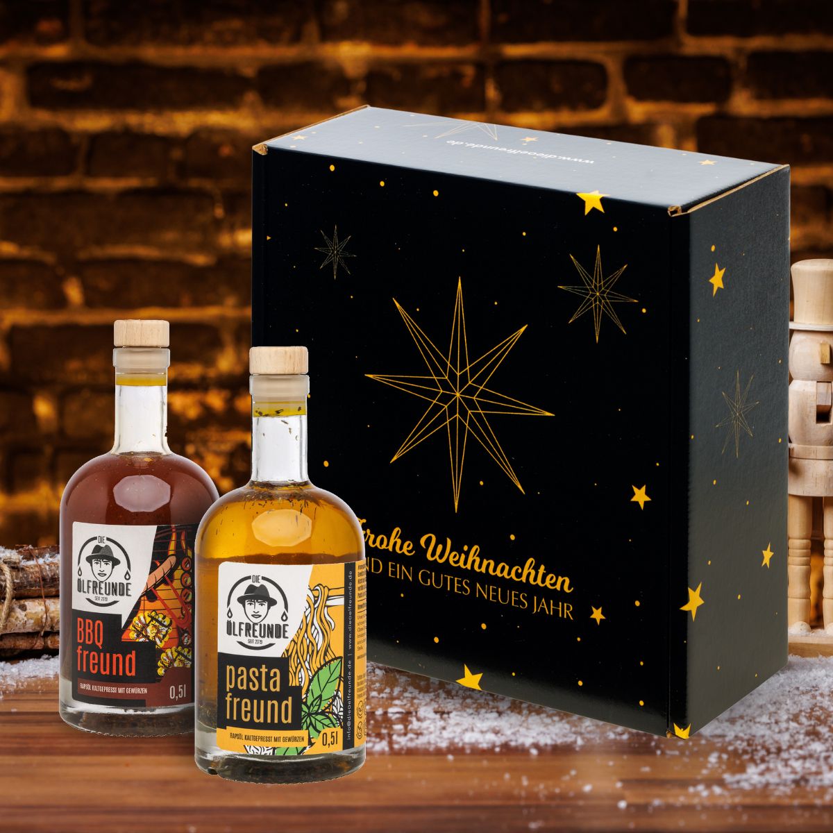 Eine Flasche BBQ-Öl und eine Flasche Pasta-Öl der Marke die Ölfreunde vor einer Weihnachts-Geschenkverpackung, neben einem Bündel Holz.