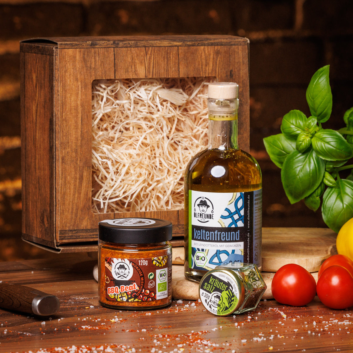 Eine Flasche Leindotteröl mit Gewürzen, ein Glas BBQ-Beat Gewürzmischung und ein Glas Kräutersalz der Marke Die Ölfreunde, vor einer Geschenkverpackung, auf einem holzigen Untergrund, neben frischem Gemüse