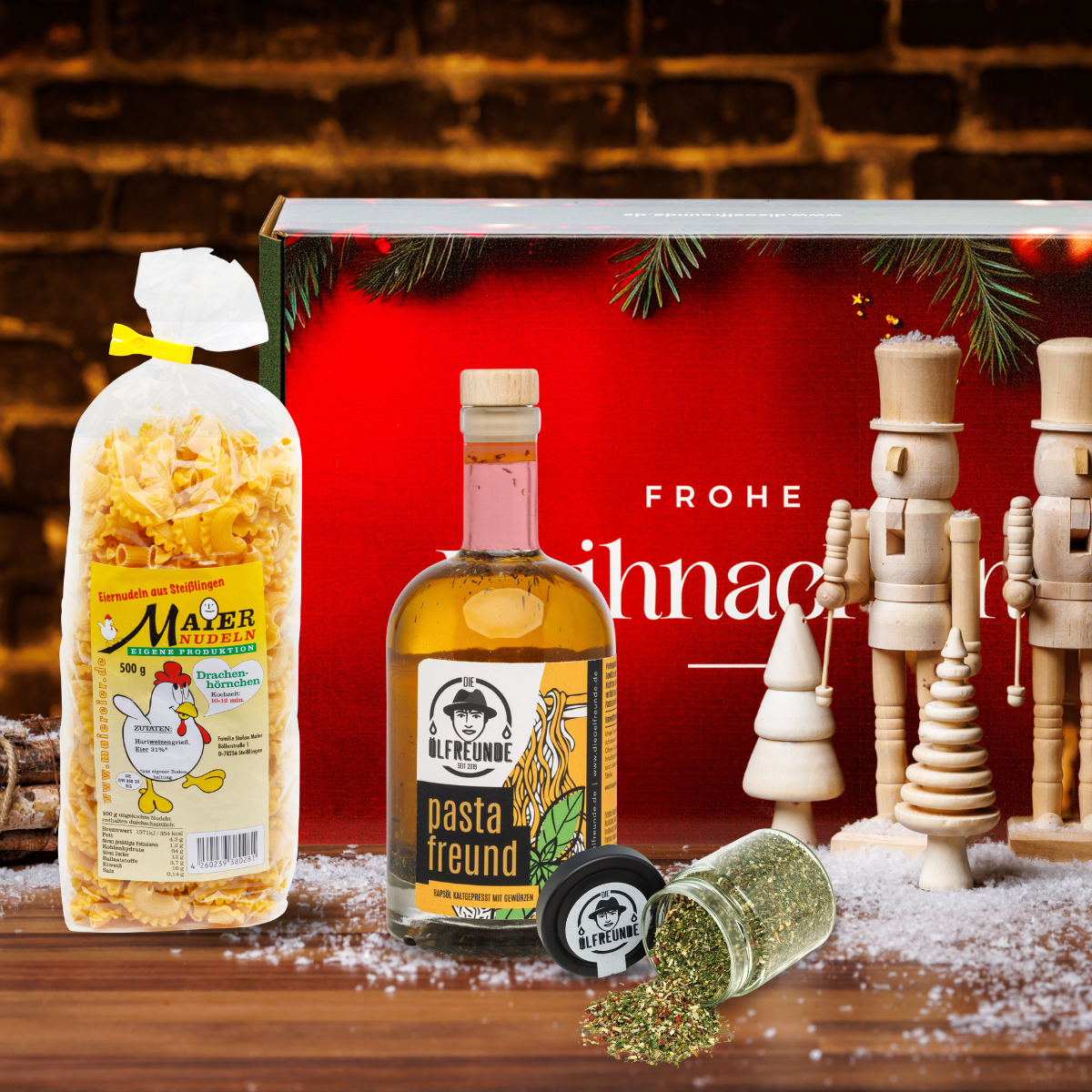 Eine Packung Drachenhörnchen-Nudeln der Marke Maier Nudeln, eine Flasche Pasta-Öl und ein Glas mit einer Gewürzmischung der Marke Die Ölfreunde, vor einer Weihnachts-Geschenkverpackung, neben weihnachtlicher Dekoration.