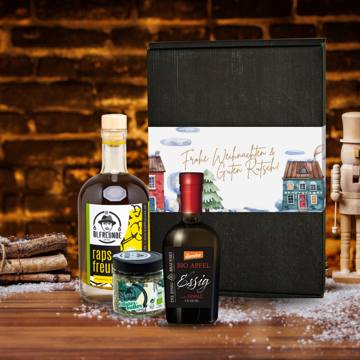 Eine Flasche Bio-Apfelessig der Marke Die Essig Brauerei, Eine Flasche Rapsöl und ein Glas Kräuterbutter-Gewürzmischung der Marke Die Ölfreunde, vor einer Weihnachts-Geschenkverpackung, auf einem holzigen Untergrund.
