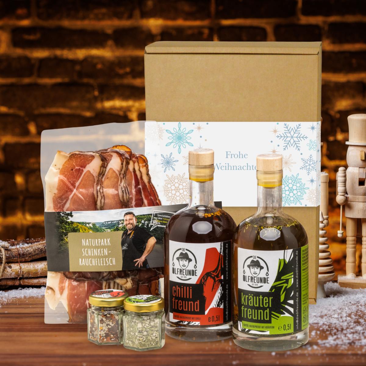 Kräuteröl, Chilli-Öl, Kräutersalz und Chillisalz der Marke Die Ölfreunde sowie Schinken-Rauchfleisch der Marke Naturpark vor einer Weihnachts-Geschenkverpackung und dunklem Hintergrund, neben einem Bündel Holz.