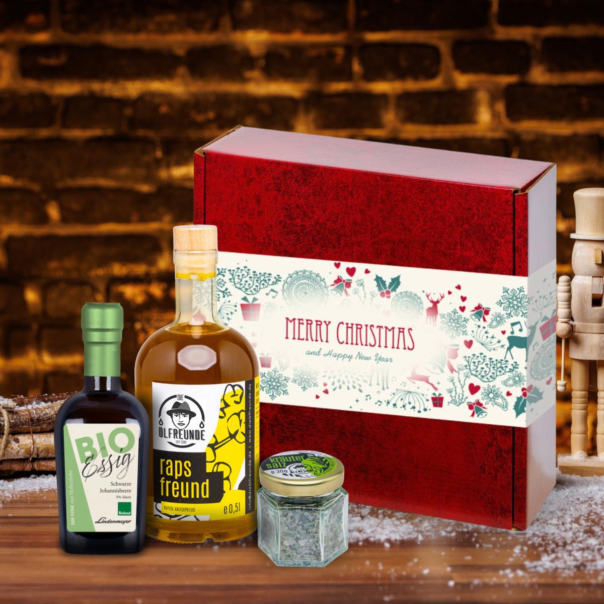 Rapsöl, Kräutersalz und Schwarze Johannisbeeren-Essig vor einer Weihnachts-Geschenkverpackung, neben einem Bündel Holz.
