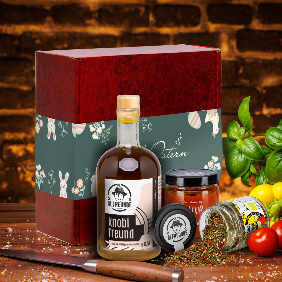 Knoblauchöl und Rühreigewürz der Marke Die Ölfreunde sowie Tomatensoße Natur der Marke Hagdorn Tomaten vor einer Oster-Geschenkverpackung, daneben ein Messer, frische Paprika, frischer Basilikum und frische Tomaten.