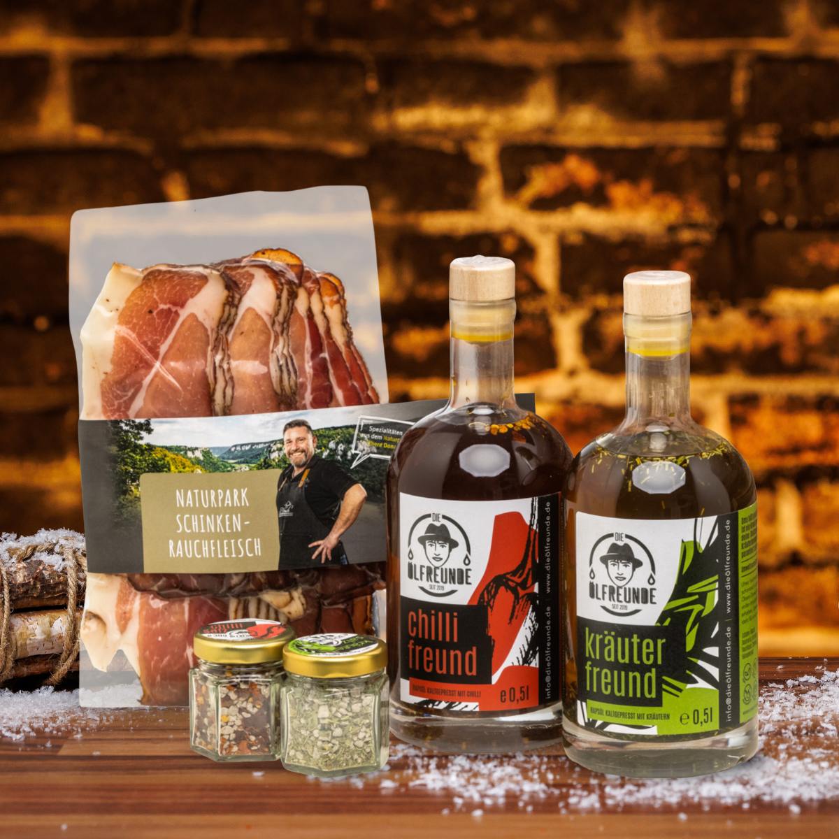 Kräuteröl, Chilli-Öl, Kräutersalz und Chillisalz der Marke Die Ölfreunde sowie Schinken-Rauchfleisch der Marke Naturpark vor dunklem Hintergrund, neben einem Bündel Holz.