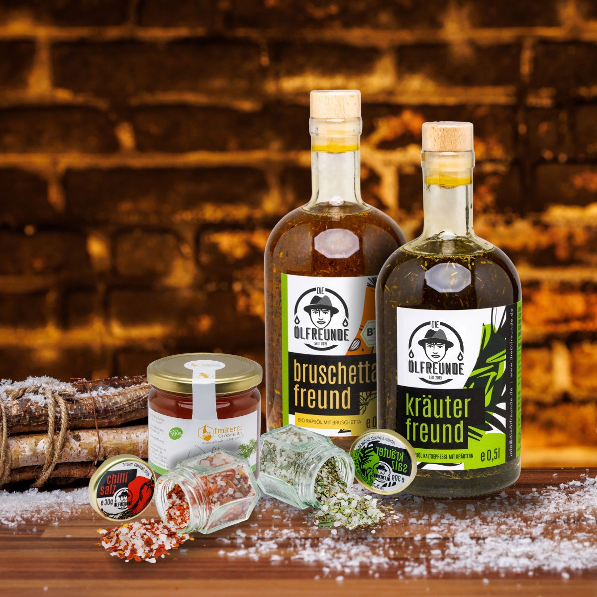 Bruschettaöl, Kräuteröl, Kräutersalz und Chillisalz der Marke Die Ölfreunde sowie Waldhonig der Imkerei Großmann vor dunklem Hintergrund, neben einem Bündel Holz.