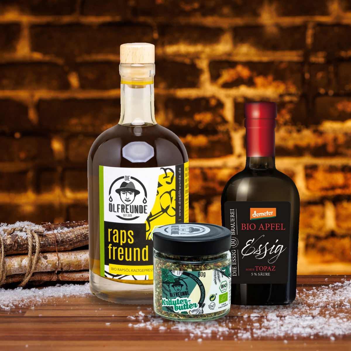 Rapsöl und Kräuterbutter Gewürz der Marke Die Ölfreunde und Bio Apfelessig der Marke Demeter, vor einem dunklen Hintergrund, neben einem Bündel Holz.