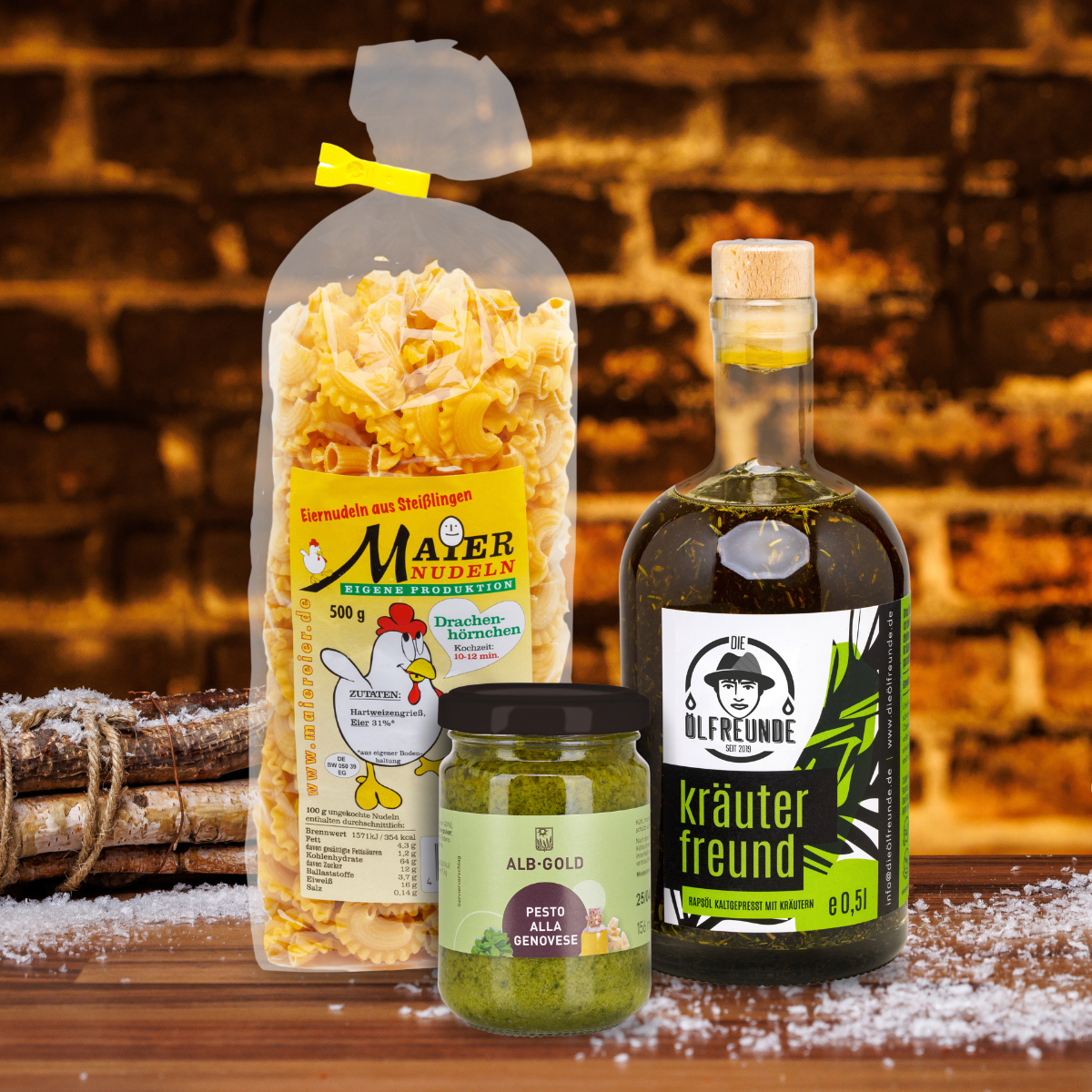 Kräuteröl der Marke Die Ölfreunde sowie Eiernudeln der Marke Maier Nudeln und Pesto alla Genovese der Marke Alb-Gold vor dunklem Hintergrund, neben einem Bündel Holz.