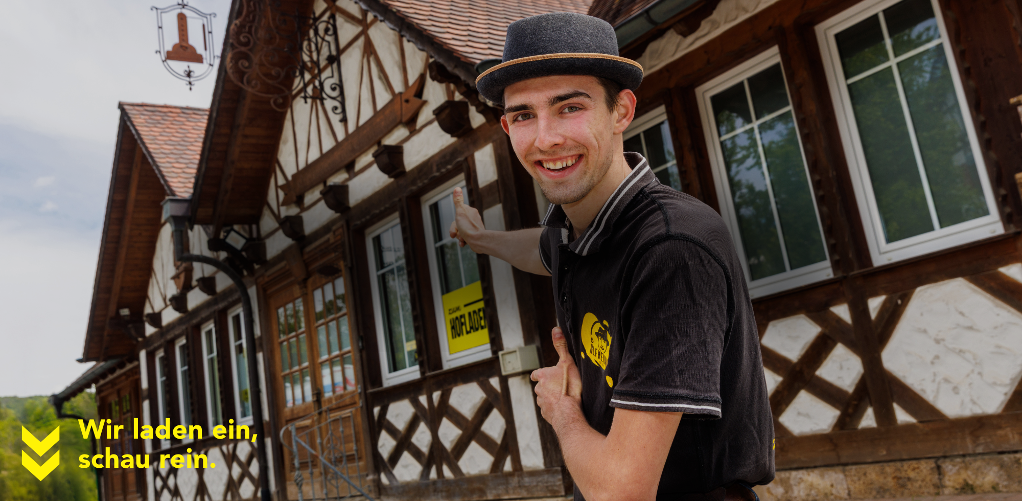 Paul vor der Ölmühle – Einladung zum Besuch mit gelbem Schild und Fachwerkhaus im Hintergrund. Persönlich, lokal und charmant – mit dem Slogan ‚Wir laden ein, schau rein