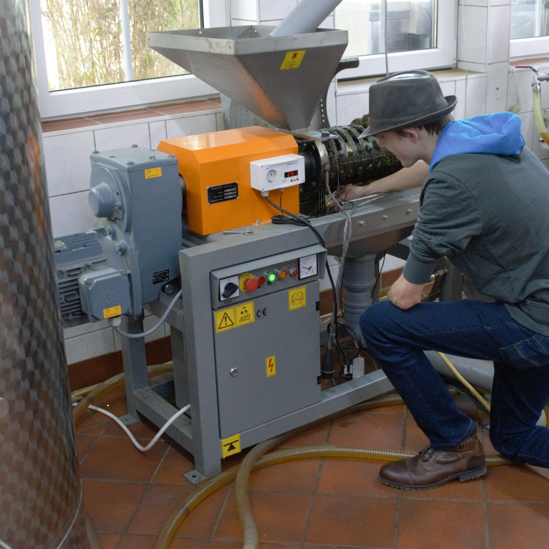 Eine Person mit Hoodie und Hut, in einer Produktion, die kniend an einer Maschine arbeitet.