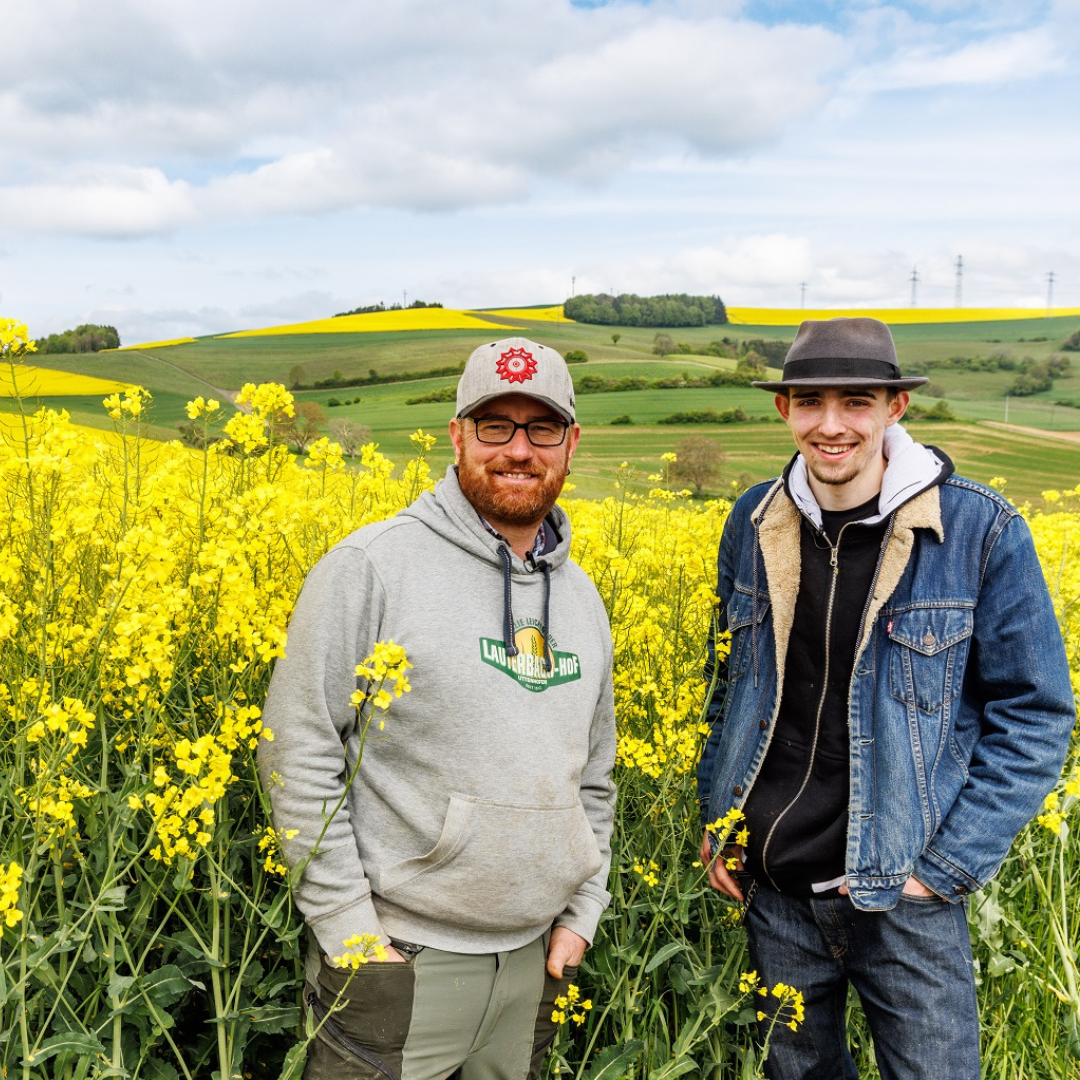 Eine Person mit grauer Cap und grauem Hoodie, daneben eine Person mit schwarzen Hut, schwarzem Hoodie und Jeansjacke vor einem Rapsfeld.