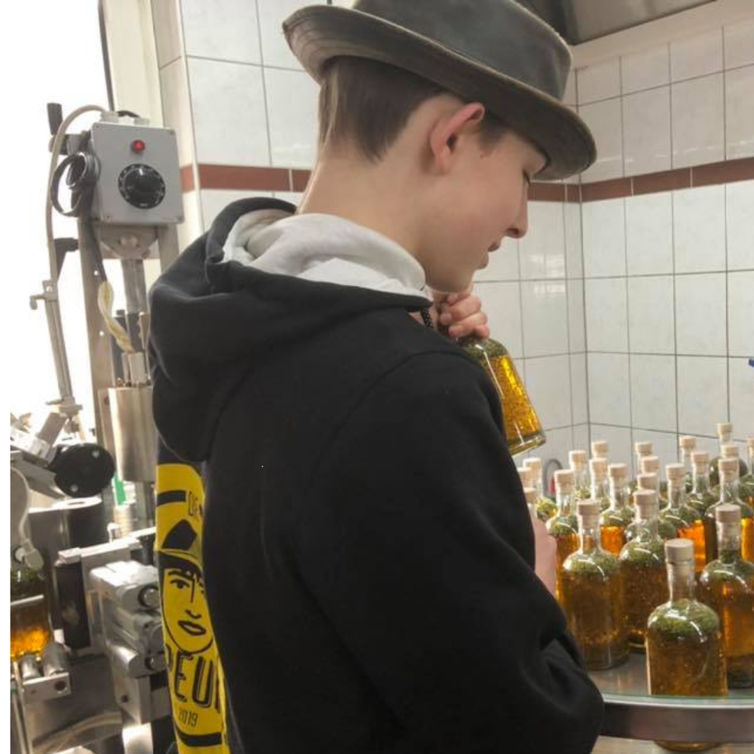 Eine Person in einem schwarzen Hoodie mit gelbem Logo der Ölfreunde auf dem Rücken und einem Hut. Die Person steht in einer industriellen Produktions- oder Abfüllanlage, hält eine Flasche mit gelben Öl in der Hand und inspiziert sie. Im Vordergrund sind mehrere ähnliche Flaschen mit Korkverschlüssen auf einem Tisch