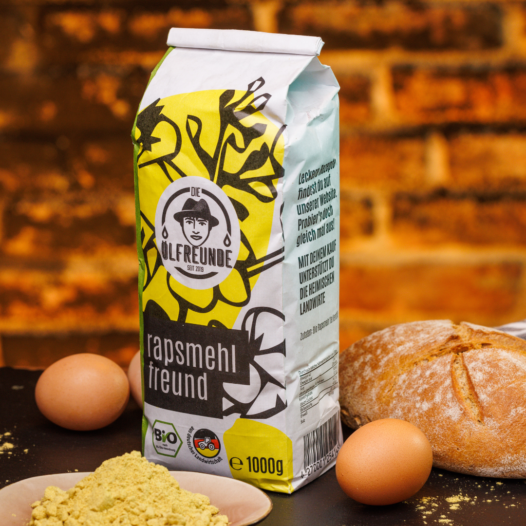 Eine Tüte mit 1000g Rapsmehl der Marke Die Ölfreunde, umgeben von Eiern und einem Frischen Brot. Davor steht eine kleine Schüssel mit Rapsmehl.