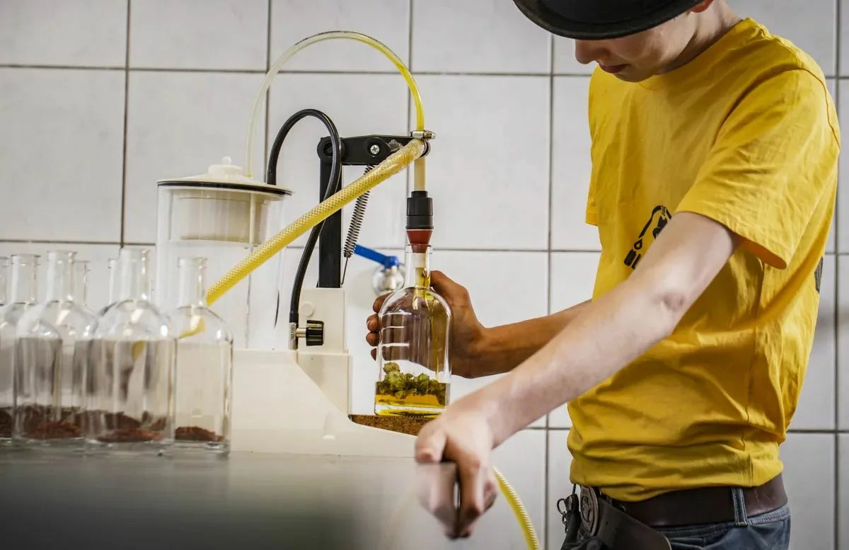 Eine Person mit schwarzen Hut und gelben T-Shirt, die dabei ist Öl in eine Flasche abzufüllen.