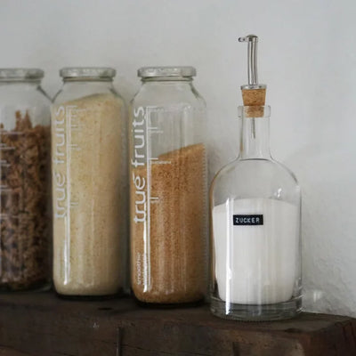 vier Glasbehälter, die ordentlich auf einem hölzernen Regal angeordnet sind. Drei der Behälter tragen das Label 'true fruits' und enthalten unterschiedliche Getreidearten oder Pulver, während der vierte Behälter mit 'ZUCKER' beschriftet ist und weißen Kristallzucker enthält. Der 'ZUCKER'-Behälter verfügt zudem über einen Korkverschluss