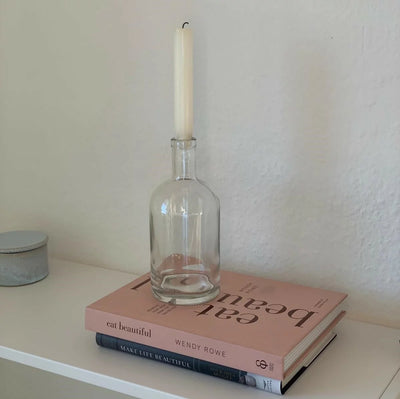 Eine Anordnung auf einem weißen Regal: Zwei übereinander gestapelte Bücher ein rosa farbiges und ein dunkel farbiges. Auf den Büchern steht ein klares Glas, in dessen Hals eine weiße Kerze eingesetzt ist. Links neben den Büchern befindet sich ein kleiner, runder, hellgrauer Behälter mit Deckel.