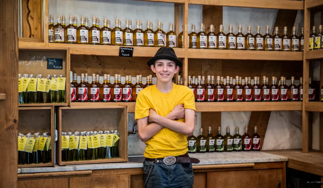 Eine Person mit schwarzem Hut und gelben T-Shirt, die mit verschränkten Armen vor einem Regal voller Öl von der Marke Die Ölfreunde steht.