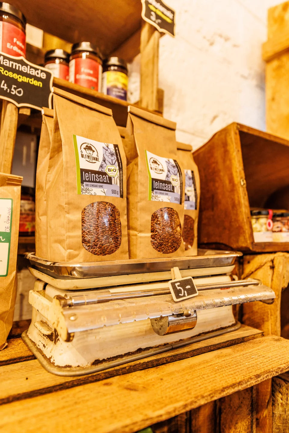 Eine historische Waage mit mehreren Packungen Bio-Leinsaat der Marke Die Ölfreunde. Darunter ist ein Preisschild in schwarz zu sehen und über den Leinsaaten stehen auch andere Produkte.