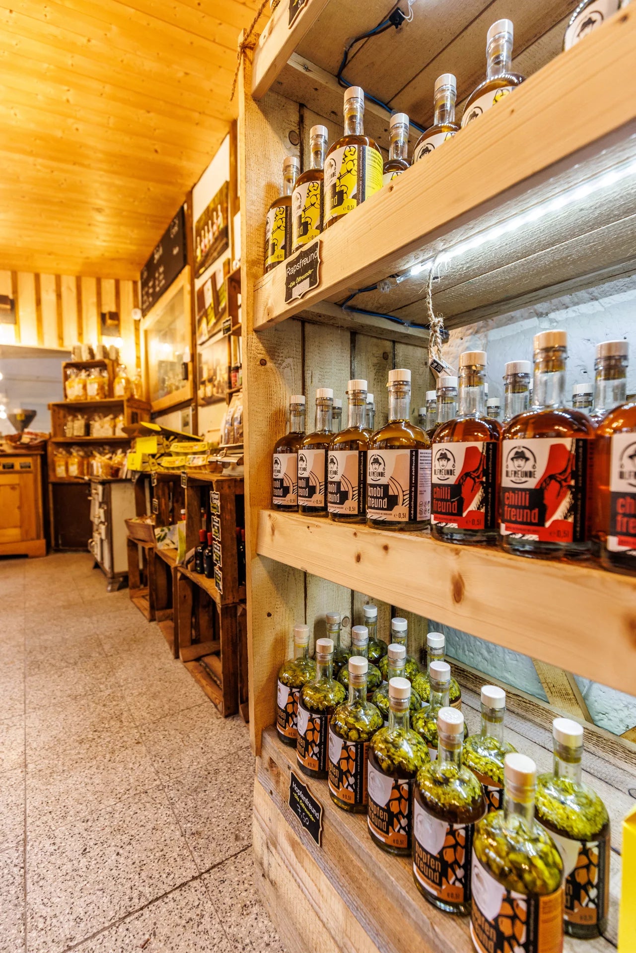 Das Innere eines Lebensmittelladens. Üppige, hölzerne Regale sind gefüllt mit Flaschen, Gläsern, Gläsern und verschiedenen Lebensmittelverpackungen. An den Wänden befinden sich diverse Schilder und Bilder sowie kleinere Hinweise zu Produkten und Preisen.