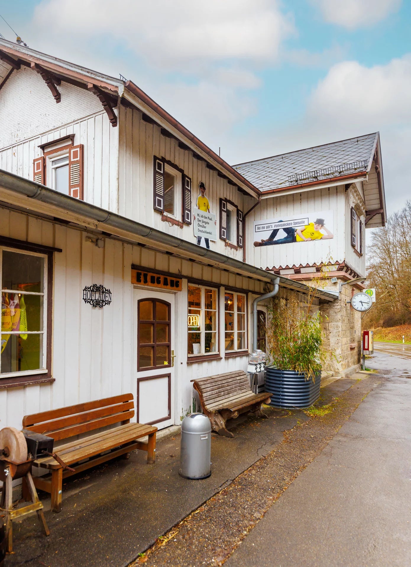 Ein traditionelles Deutsches Gebäude. Davor stehen zwei Holzbänke und ein Mülleimer. Am Haus sind mehrere Schilder angebracht, die auf den Hofladen verweisen oder Personen abbilden. Außerdem ist an dem Gebäude eine alte Bahnhofsuhr und ein Haltestellenzeichen zu sehen.