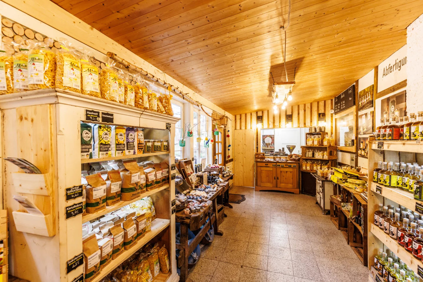 Das Innere eines Lebensmittelladens mit rustikalem Charme. Üppige, hölzerne Regale sind gefüllt mit Flaschen, Gläsern, Gläsern und verschiedenen Lebensmittelverpackungen. Mehrere warm leuchtende Glühbirnen, die von der Decke hängen. An den Wänden befinden sich diverse Schilder und Bilder – darunter ein Schild mit den Aufschrift 'Abfertigung' sowie kleinere Hinweise zu Produktnamen und Preisen.