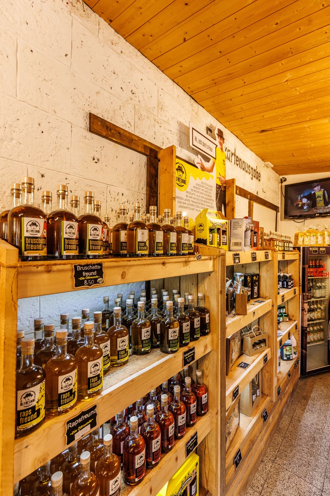 Das Innere eines Lebensmittelladens. Üppige, hölzerne Regale sind gefüllt mit Flaschen, Gläsern, Gläsern und verschiedenen Lebensmittelverpackungen. An den Wänden befinden sich diverse Schilder und Bilder sowie kleinere Hinweise zu Produkten und Preisen.