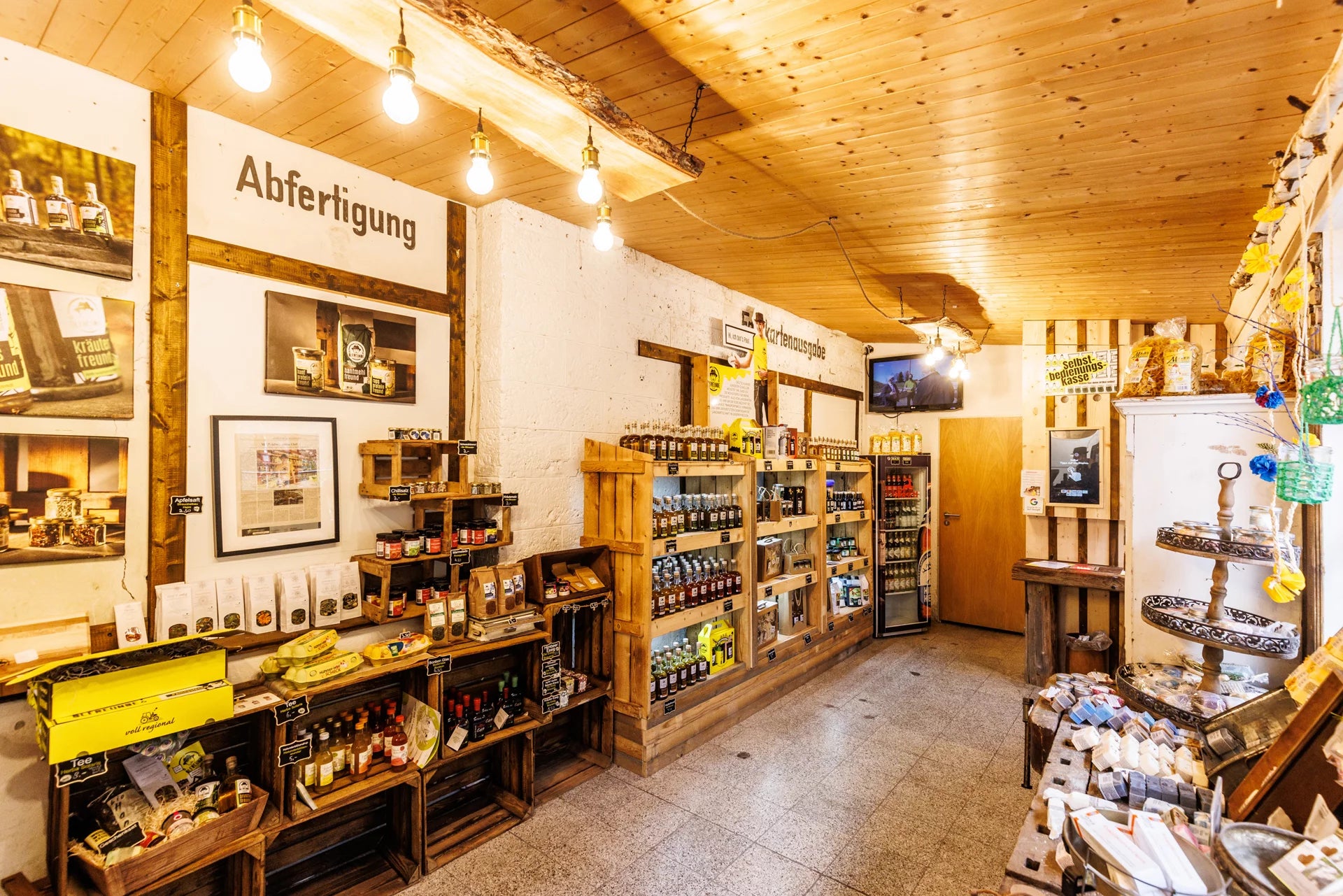 Das Innere eines Lebensmittelladens mit rustikalem Charme. Üppige, hölzerne Regale sind gefüllt mit Flaschen, Gläsern, Gläsern und verschiedenen Lebensmittelverpackungen. Mehrere warm leuchtende Glühbirnen, die von der Decke hängen. An den Wänden befinden sich diverse Schilder und Bilder – darunter ein Schild mit den Aufschrift 'Abfertigung' sowie kleinere Hinweise zu Produktnamen und Preisen.