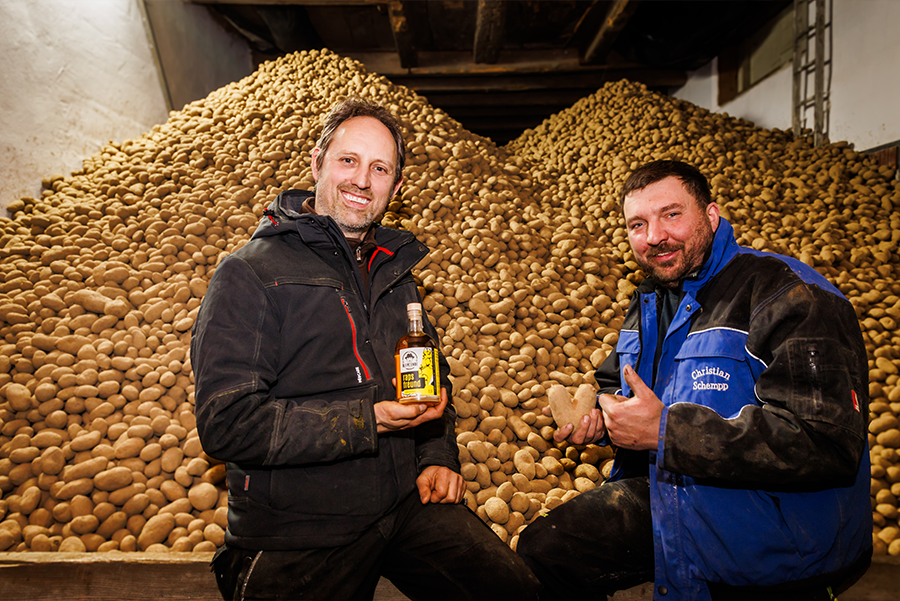 2 Männer sitzen vor einem riesen Kartoffelberg. Einer der beiden Männer hat eine graue Jacke an und eine Ölflasche mit gelben Etikett in der Hand. Der andere trägt eine blaue Jacke und hält eine herzförmige Kartoffel in der Hand während er einen Daumen nach oben gibt.
